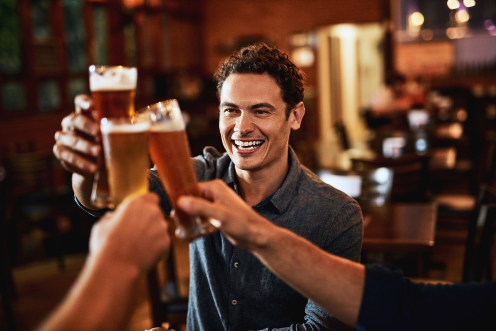 A group of young friends seated at a table together while enjoying a beer and celebrating with a toast.