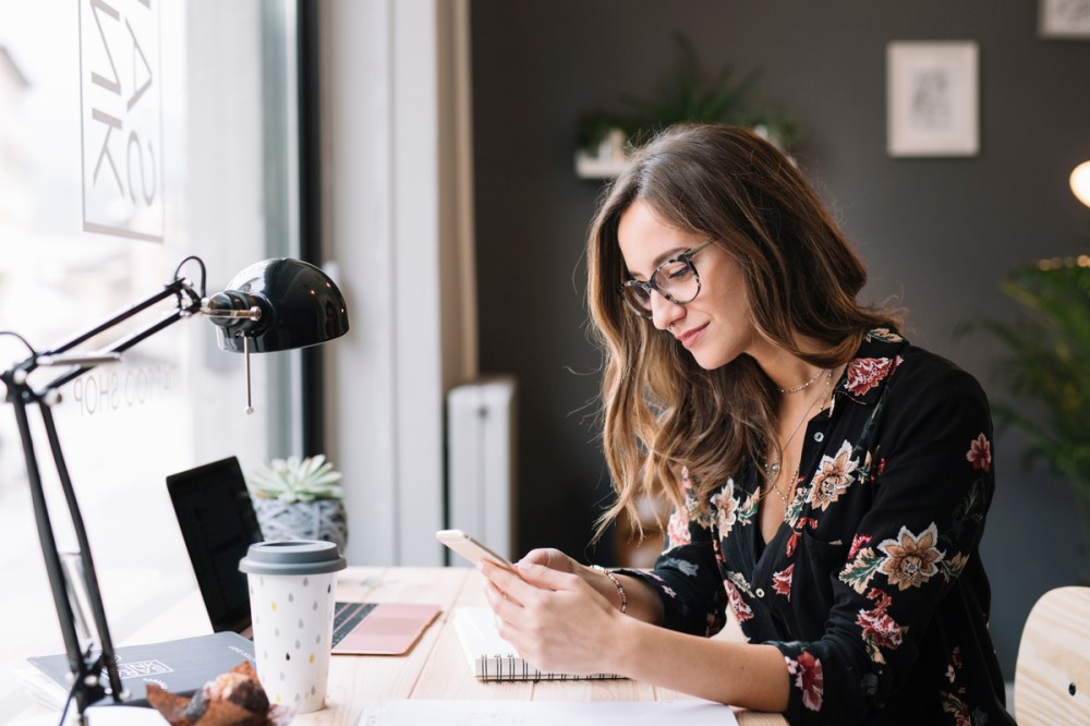A woman wearing a floral print is sitting at a desk with a laptop, looking at a phone in her hand.