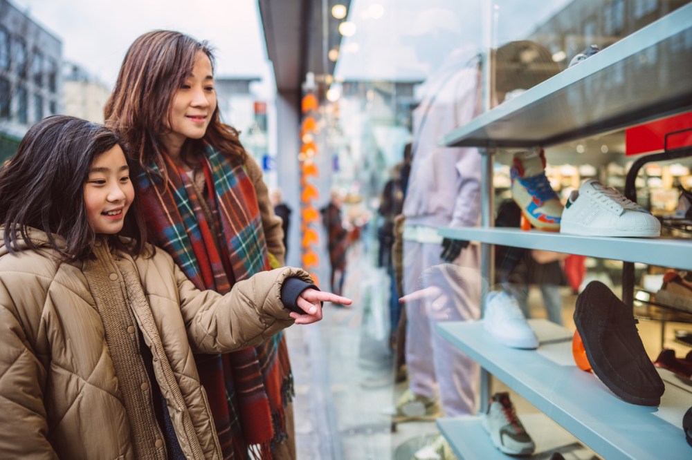 girl looking at window display with her young pretty mom while walking on shopping street.