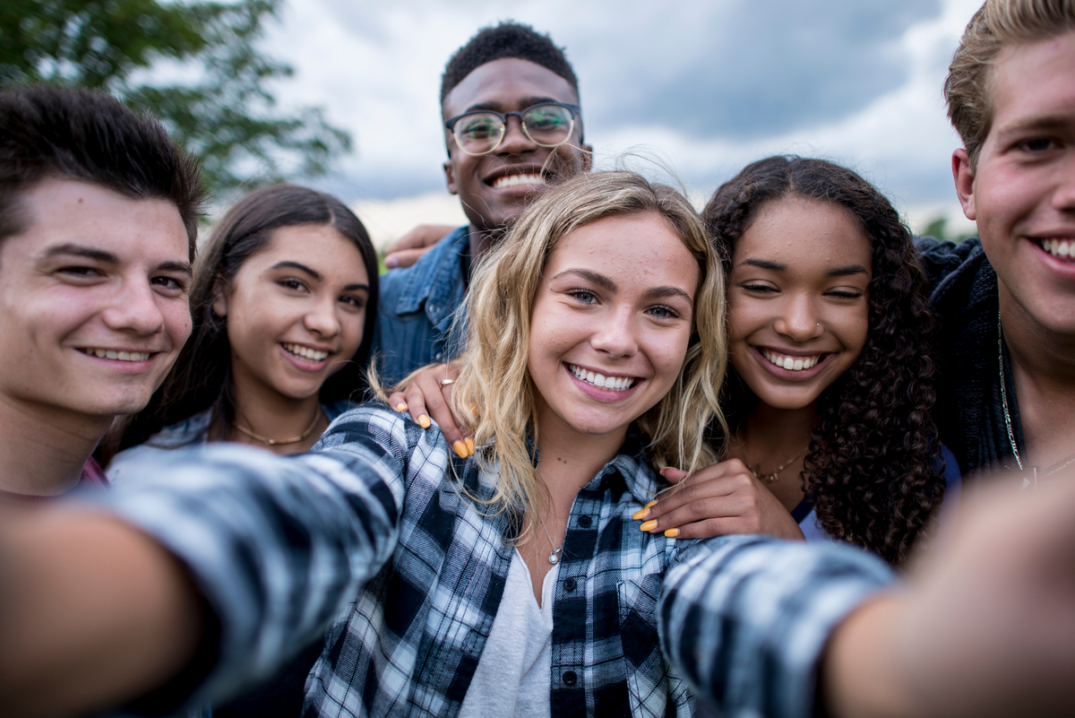 A multi-ethnic group of teenagers are outdoors on a cloudy day. They are wearing casual clothing. They are smiling while taking a selfie together.