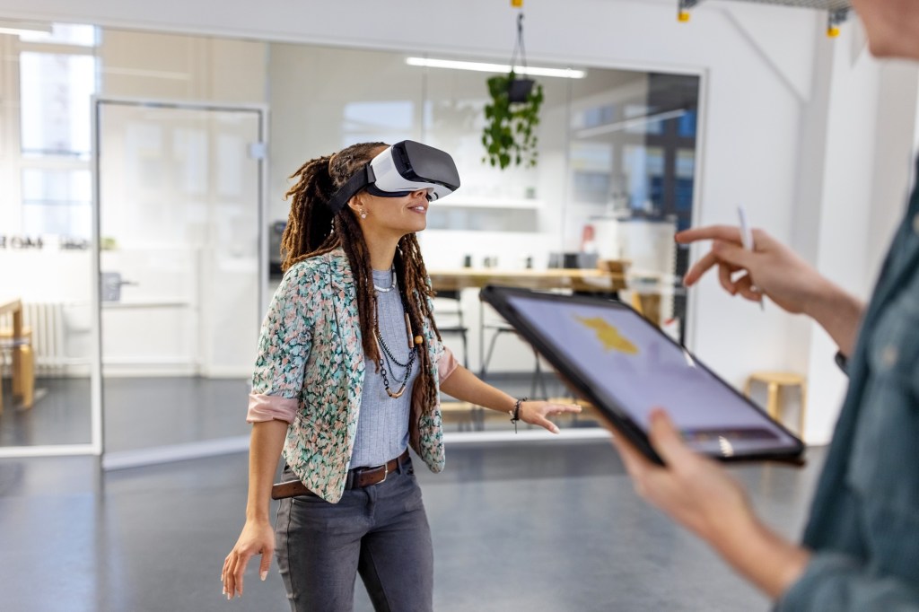Young woman wearing virtual reality headset by a male colleague using digital tablet. Startup business colleagues testing out virtual reality glasses in the office.