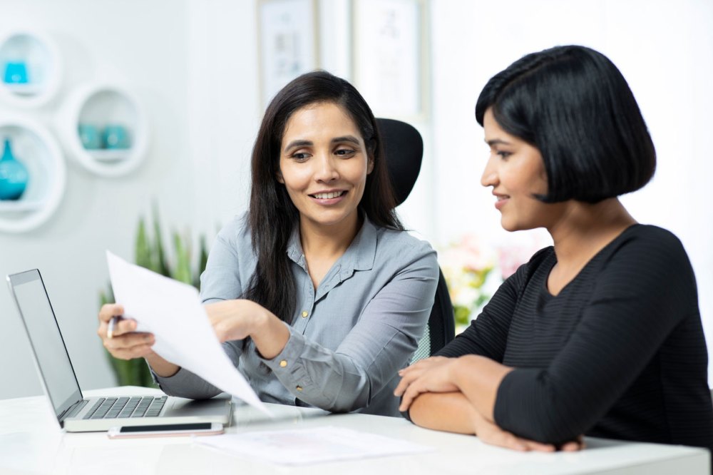 Two women look over a paper near a laptop in a bright room.