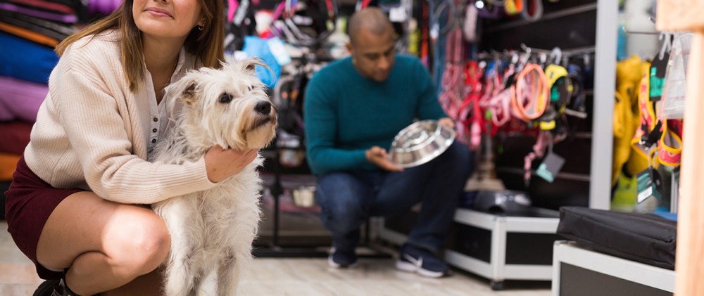 A woman pets a white dog while a man looks at a dog bowl in a pet store.