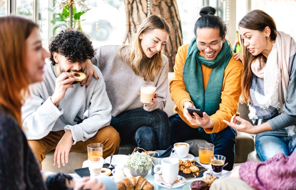 Multicultural friends playing with mobile phone at coffee bar - People having fun together at cafeteria on brunch time - Life style concept with happy men and women at cafe venue - Bright warm filter