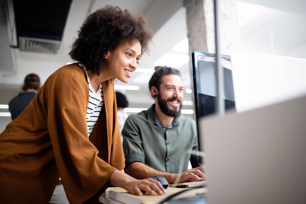 A women with an orange shirt works with a bearded man with a green shirt in an office setting