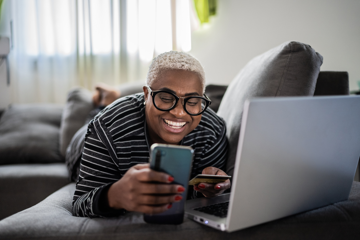 Woman in a striped shirt wearing thick framed glasses holding a smart phone and a credit card making a purchase on her laptop while lying on a couch