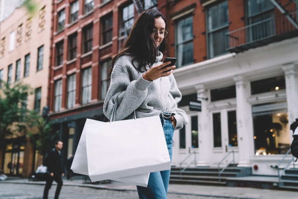 Happy slim attractive woman in glasses with shopping bags wearing denim and sweater listening to music with earphones using smartphone on city street