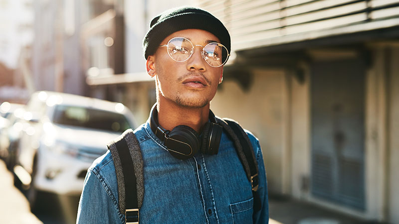 Asian man with glasses and a hat wearing headphones with a backpack and jean shirt