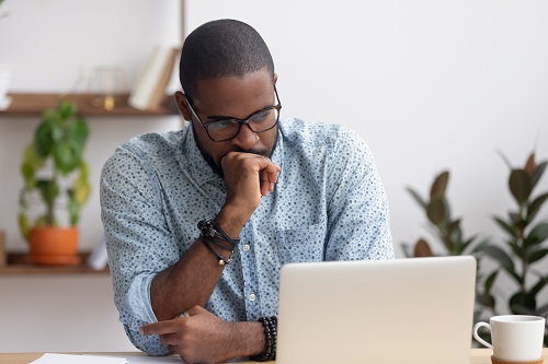 Man with glasses and light blue dress shirt looking pensive as he works on his laptop, a cup of coffee next to him with plants behind him