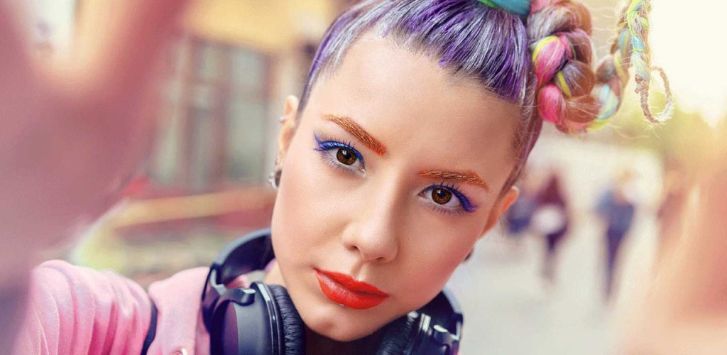 Woman with heavy makeup and colored hair with headphones and a pink hoodie taking a selfie