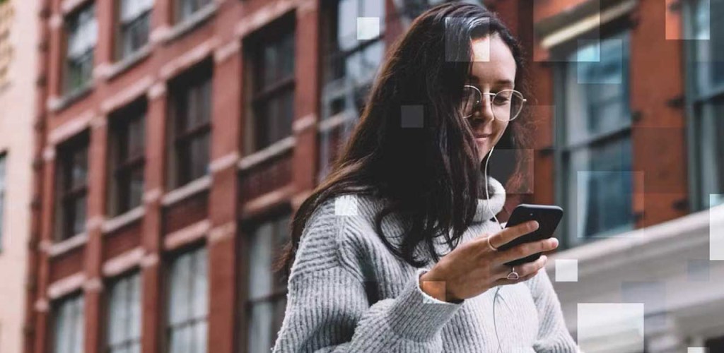 Woman with dark hair and glasses on a street with a gray sweater looking at her mobile phone