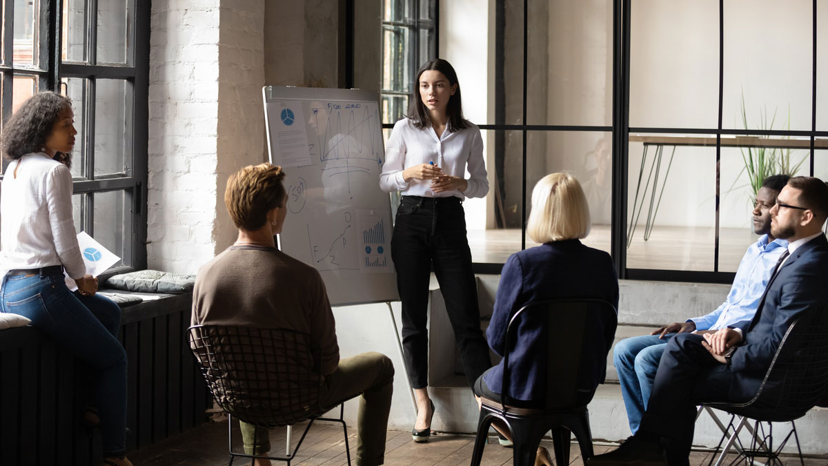 Woman with white shirt and black pants leading a meeting in a conference room with 5 seated people