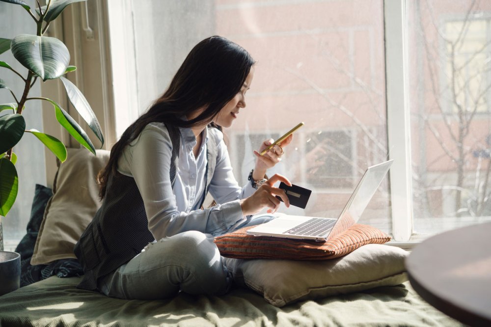 A woman with dark hair holding a credit card in one hand and a cell phone in the other with a laptop in front of her on pillows ordering something online.